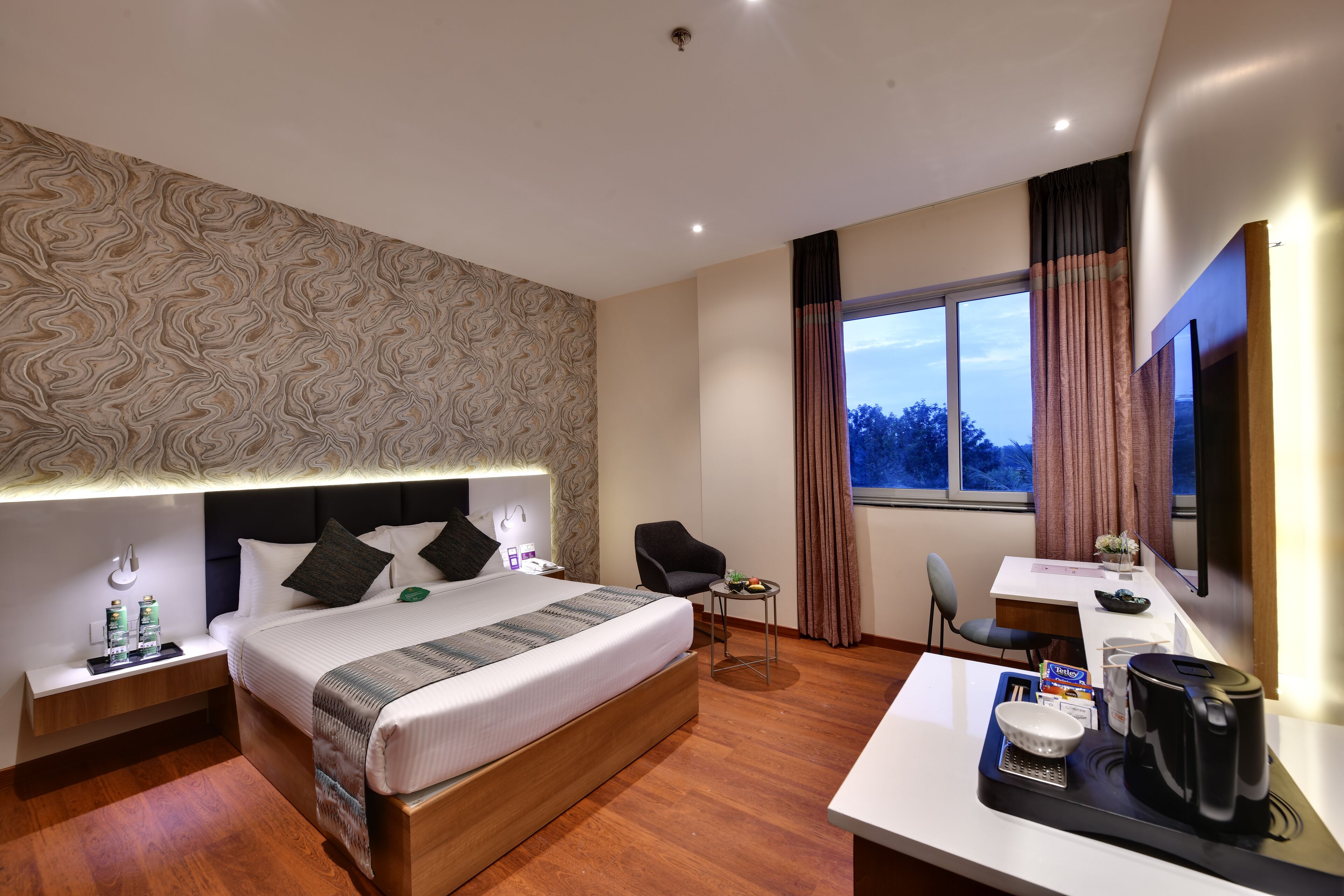 A hotel room with a large bed, a desk with a coffee maker, a chair, and a small table with a bowl and a box.