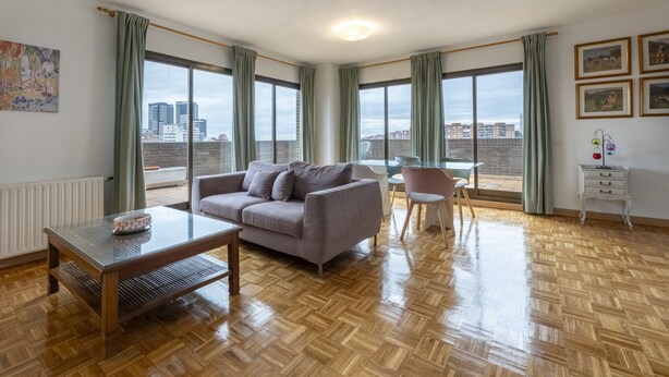 Una sala de estar moderna con un sofá, una mesa de centro y una silla. Hay ventanas grandes con cortinas, un radiador y una mesita con un objeto decorativo.
