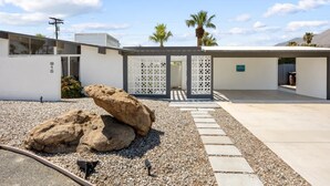 A modern building with a white facade, a large rock sculpture, and a gravel pathway leading to a gated entrance. - Mountain-View Retreat: Pet-Friendly Home with Private Pool (Palm Springs)