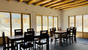 A dining area with a wooden table and chairs, large windows with a view of a building and trees, and a wooden ceiling. - Hunder Tamarix Resort (Leh)