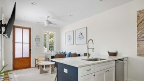 A modern kitchen with a white countertop, a built-in dishwasher, and a sink. There is a dining area with a table and chairs, and a large window with blinds.