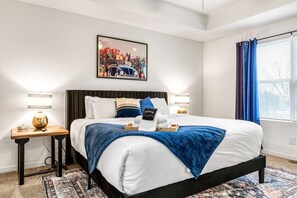Una habitación de hotel con una cama grande, una mesita de noche con lámpara, un cuadro en la pared y una ventana con cortinas.