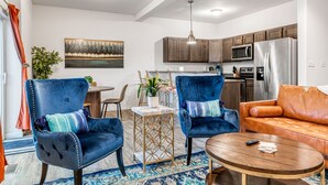 Een moderne woonkamer met twee blauwe fluweel fauteuils, een houten salontafel en een oranje leren bank. Achtergrond is een keuken met houten kastjes en roestvrijstalen apparatuur.