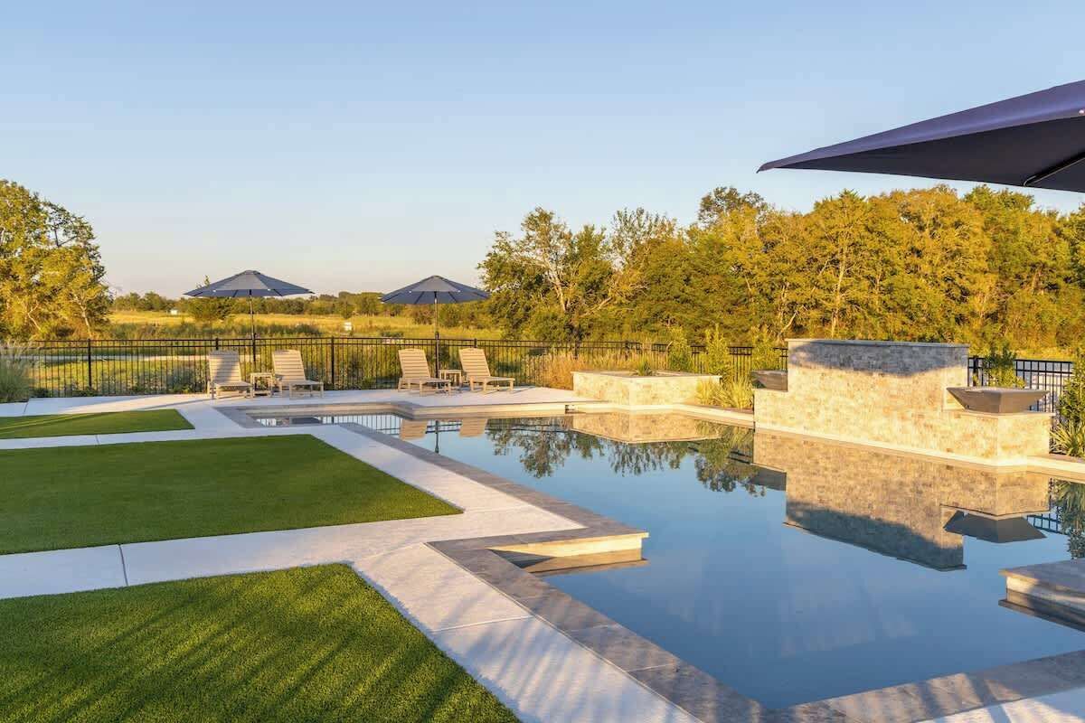 A swimming pool with lounge chairs and a stone wall, surrounded by a fence and trees.