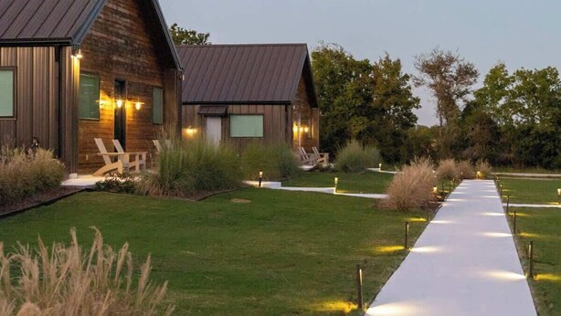 A modern building with a metal roof and wooden walls, surrounded by a well-maintained lawn and a concrete pathway leading to the entrance. - Elevated Escape | Water Retreat @ Element Ranch (Carmine)