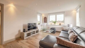 A modern living room with a flat-screen TV, leather sofa, glass coffee table, and large windows.