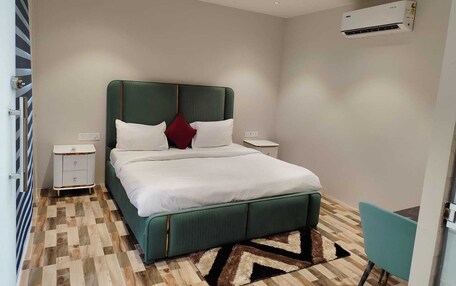 A modern hotel room with a green upholstered bed, white bedding, a small bedside table, a nightstand, a teal chair, and a patterned rug.