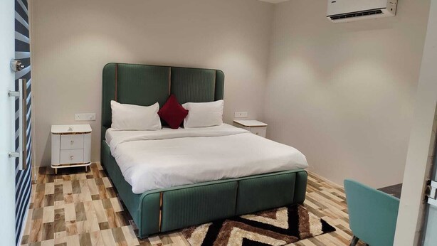 Ett modernt hotellrum med en säng klädd i grönt tyg, vita sängkläder, ett litet sängbord, ett sidobord, en turkosfärgad stol och en mönstrad matta.
