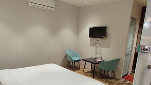 Ett hotellrum med en säng, ett skrivbord, två stolar, en tv som är monterad på väggen och en luftkonditioneringsanläggning.