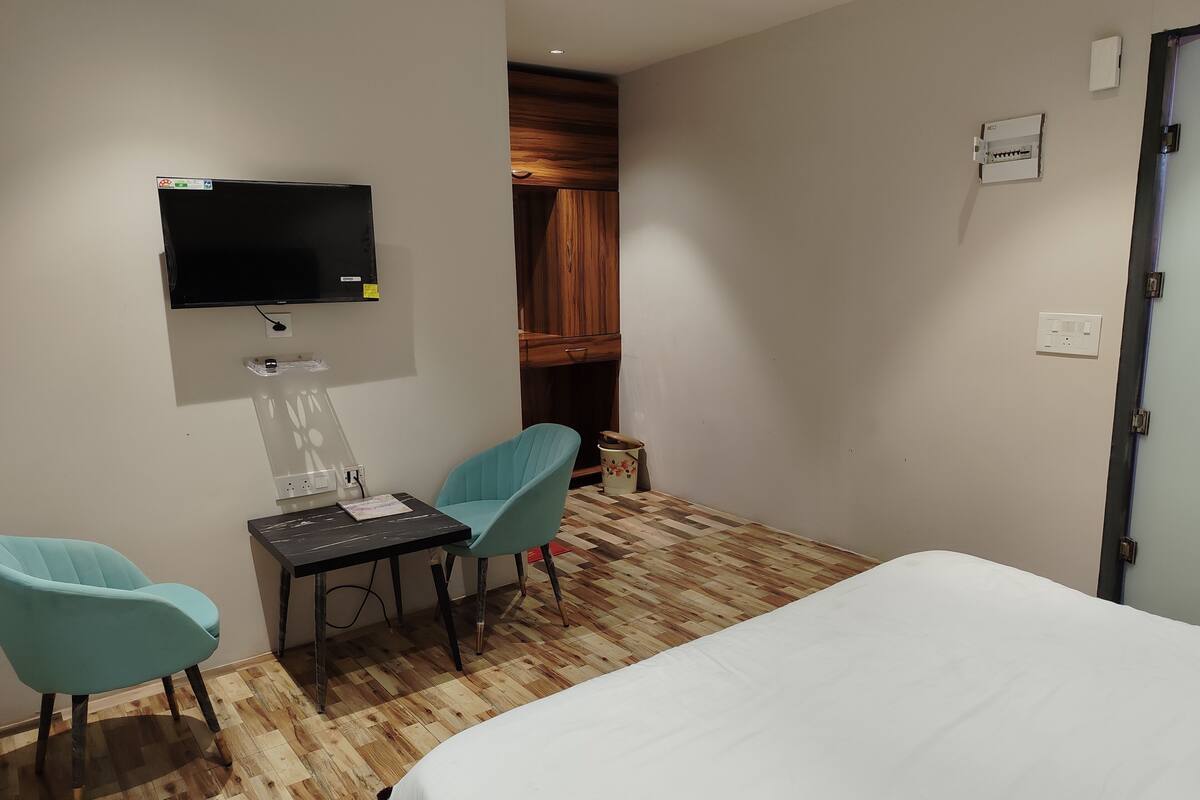 A hotel room with a bed, two chairs, a table, a TV mounted on the wall, and a wooden wardrobe.