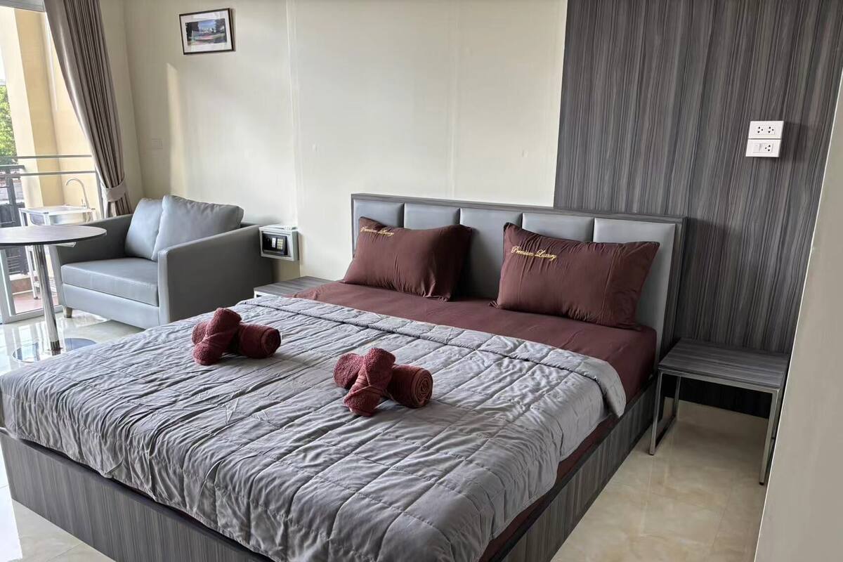 A modern hotel room with a large bed, grey and brown bedding, a grey sofa, a small table, and a bedside table with a lamp.