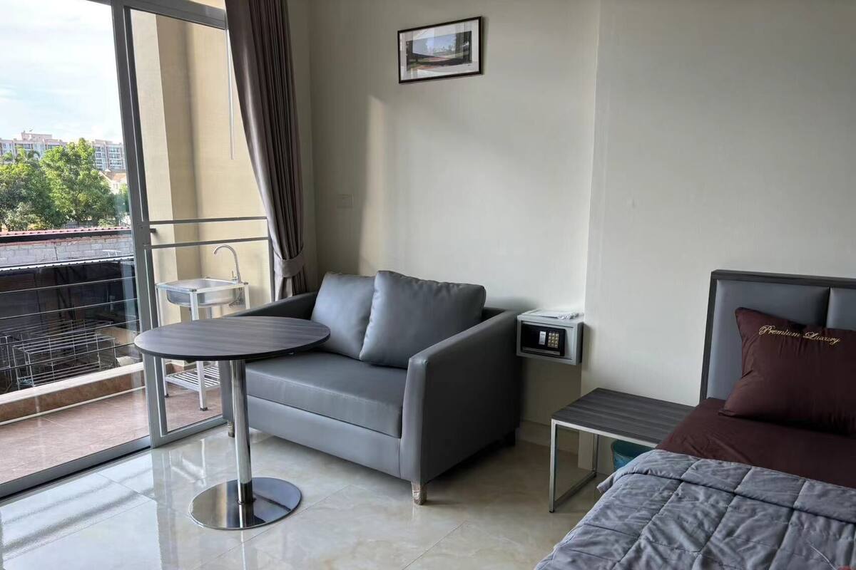 A modern hotel room with a grey sofa, a round table, a bed with a grey quilt, a small bedside table, and a view of the city through large windows.
