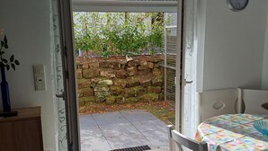 An open door leading to a patio with a stone wall, a table with a colorful tablecloth, and a clock on the wall. - Vacation apartment in the forest/ central location/ leisure facilities (Neunkirchen)