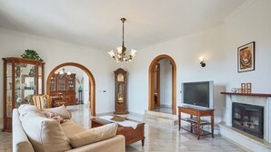 A spacious living room with a beige sofa, wooden coffee table, and a television. There is a fireplace with a mantel and a clock on the wall. - Puig de Sa Figuera Country House with Private Pool and Panoramic Views for 8 pax (Felanitx)