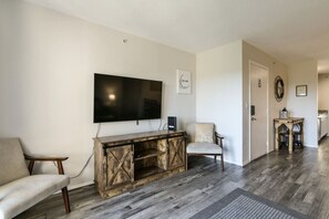 Una sala de estar con un televisor de pantalla plana grande montado en la pared, un mueble de madera para entretenimiento, dos sillones y una puerta que da a otra habitación.