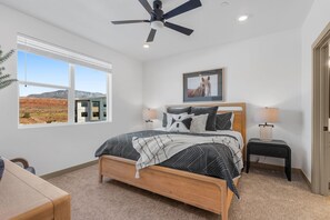 A bedroom with a large bed, two bedside tables, a ceiling fan, and a window with a view of a desert landscape. - Zion Vista Retreat | Resort Pool + Lazy River | 30 Min to Zion (Washington)