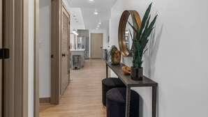 A modern hallway with a wooden floor, a console table with a mirror, a potted plant, and a door leading to a kitchen area.