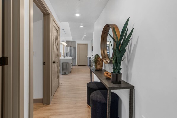 A modern hallway with a wooden floor, a console table with a mirror, a potted plant, and a door leading to a kitchen area.