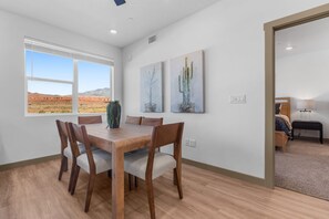 A dining area with a wooden table and chairs, a large window with a desert view, and two cactus paintings on the wall.