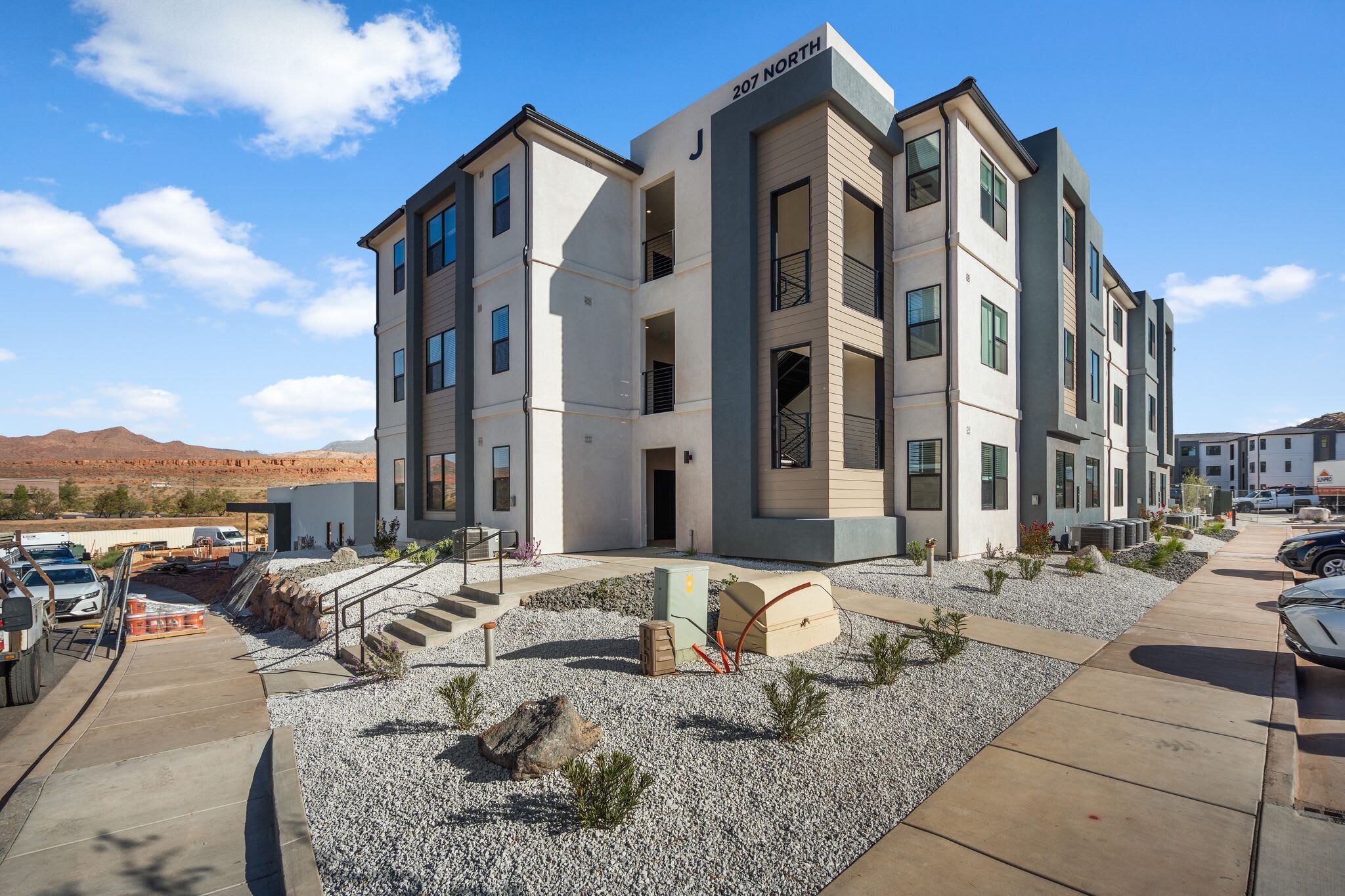 Un immeuble à plusieurs étages doté d’une architecture moderne, avec une entrée imposante agrémentée de marches et un espace paysager avec du gravier et de petites plantes.