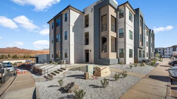 Un immeuble à plusieurs étages doté d’une architecture moderne, avec une entrée imposante agrémentée de marches et un espace paysager avec du gravier et de petites plantes.