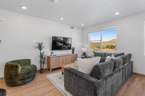 A modern living room with a large flat-screen TV, a green armchair, a grey sofa, and a view of a desert landscape through large windows.