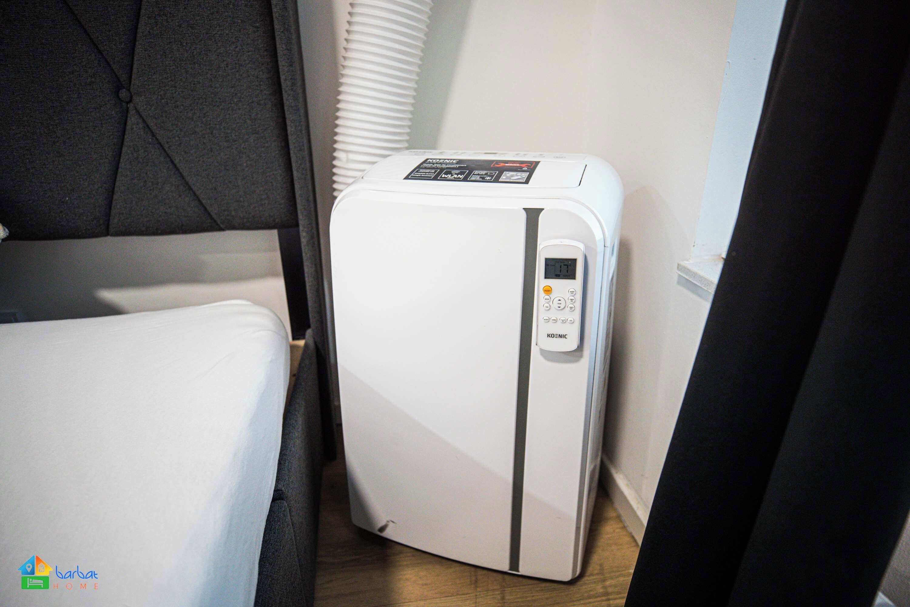 A compact white refrigerator with a digital control panel, situated in a small room with a bed and a window with blinds.