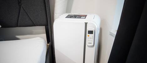 A compact white refrigerator with a digital control panel, situated in a small room with a bed and a window with blinds.