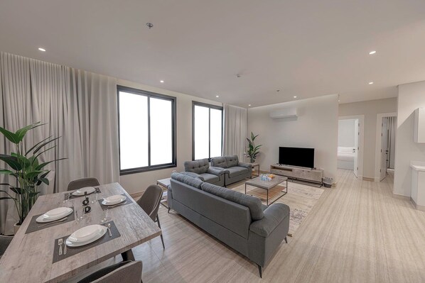 A modern living room with a grey sofa, a dining table set for four, and a flat-screen TV. - Mabaat - Al Rimal - 565 (Riyadh)