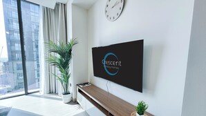 A modern living room with a large TV displaying 'Crescent Holiday Homes', a potted plant, and a view of a cityscape through large windows. - Modern 2 Bed with Stunning Views (Dubai)