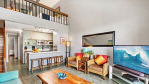 A spacious living area with a wooden coffee table, a sofa with colorful cushions, a television, and a kitchen in the background.