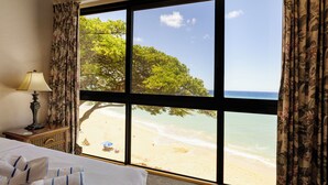 A hotel room with a large window offering a beach view, a bed with towels, and a nightstand with a lamp.