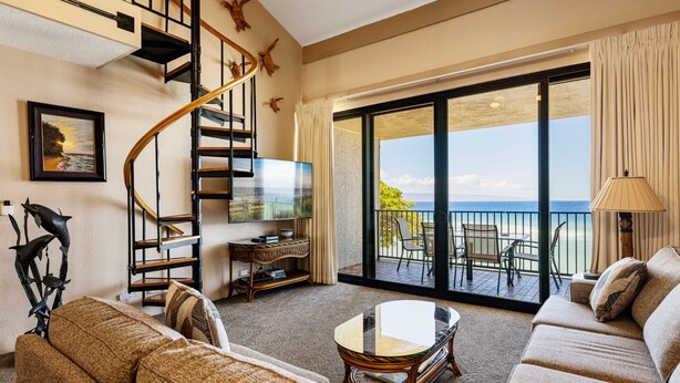 A spacious living room with a large sofa, a coffee table, a TV, and a staircase leading to a balcony with a view of the ocean.