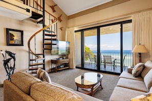 Relax in a spacious living area with a sofa set, coffee table, and wall-mounted TV. Enjoy a view of the ocean from the upper level, accessible via a sliding glass door.
