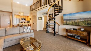 A spacious living area with a sofa, a glass coffee table, a TV, a staircase, and a kitchen in the background.