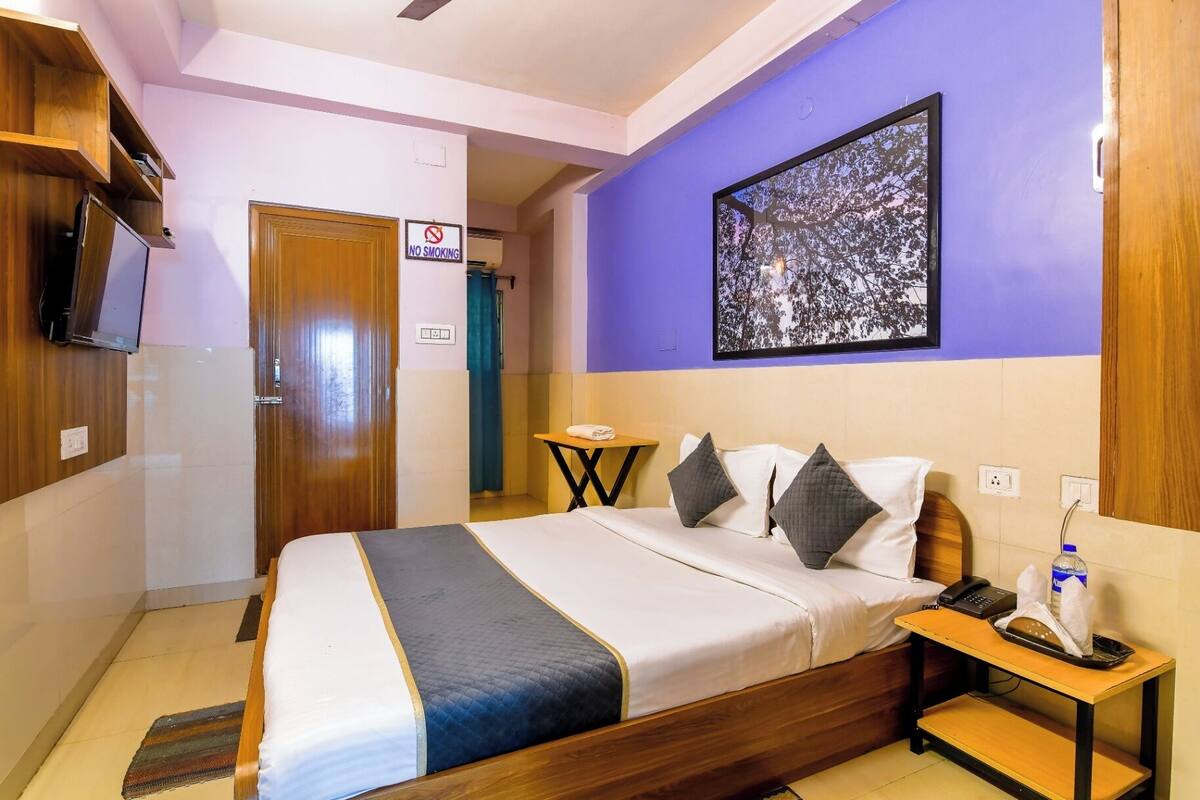 A hotel room with a bed, a bedside table with a phone and tissues, a painting on the wall, and a 'No Smoking' sign.