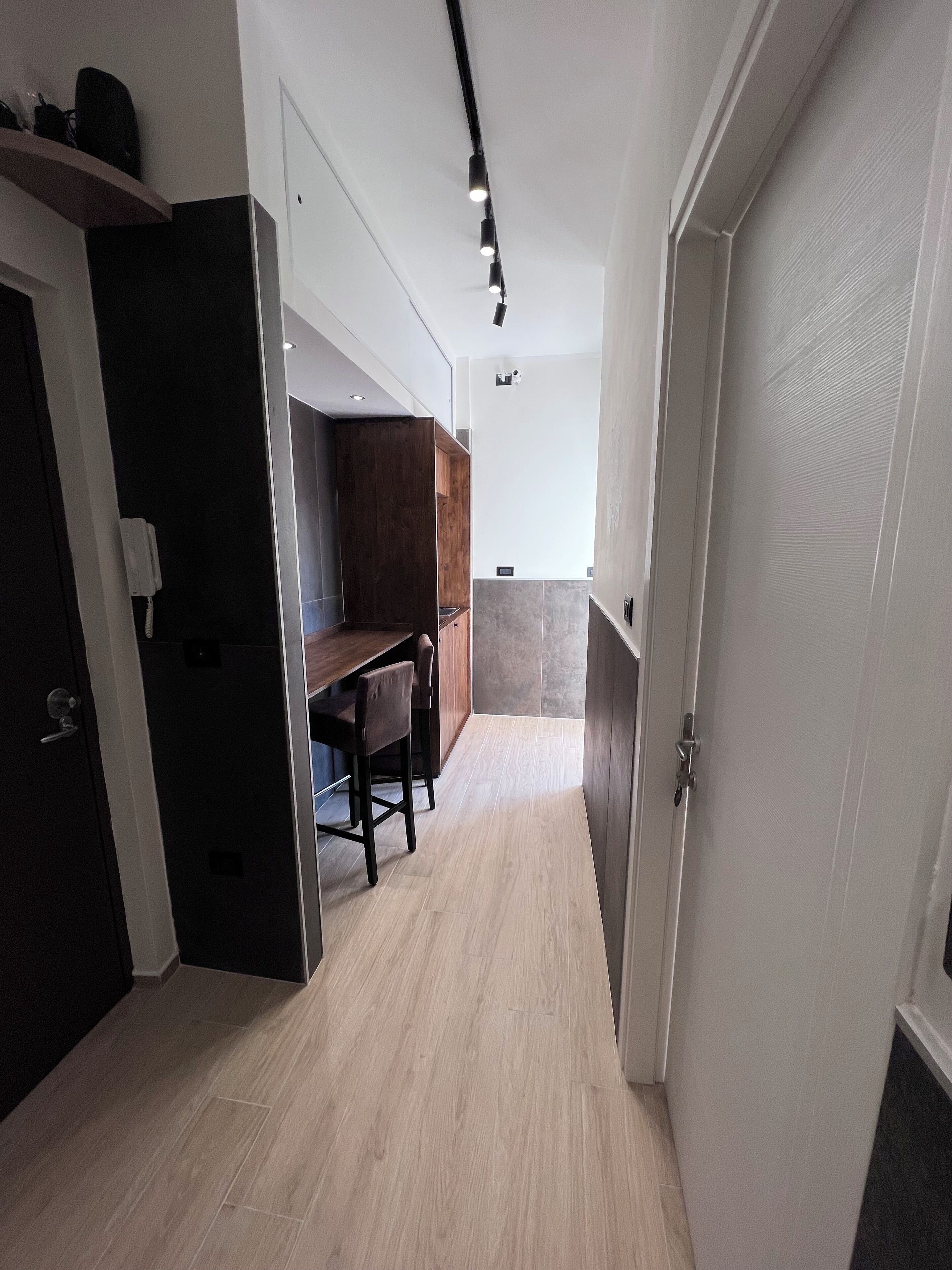 A narrow hallway with a wooden floor, a dark wooden cabinet, a small table with chairs, and a white door on the right.