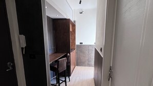 Een smalle gang met een houten vloer, een donker houten kastje, een klein tafeltje met stoelen en een witte deur aan de rechterkant.