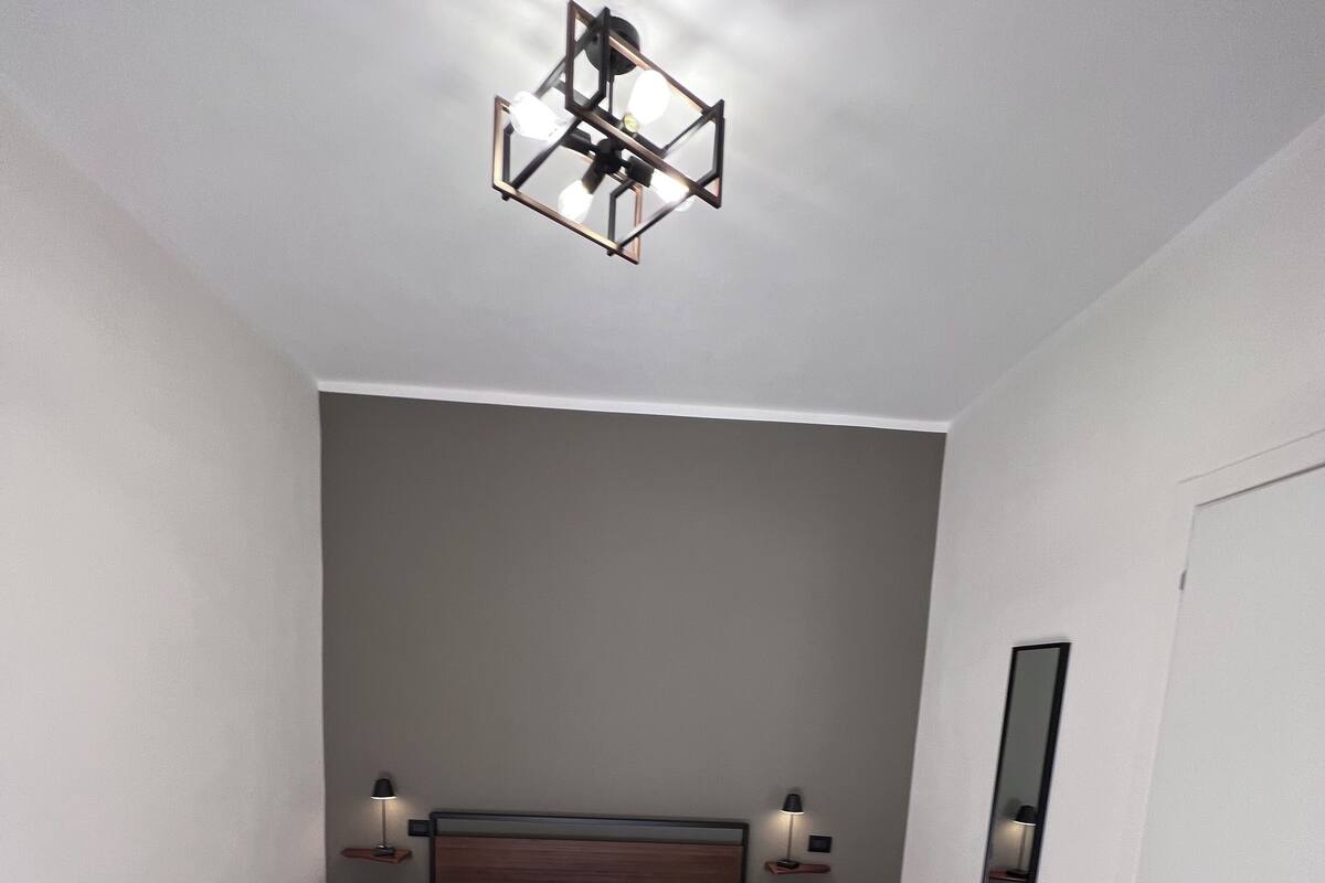 A modern bedroom with a single bed, a nightstand with a lamp, a wall-mounted mirror, and a geometric ceiling light fixture.