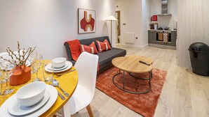 Ett kompakt bostadsutrymme med en matplats, en soffa, ett litet kök och en papperskorg.