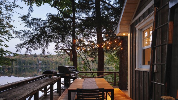 Terraza de madera con mesa y sillas, con vista a un lago, una parrilla y luces colgantes.