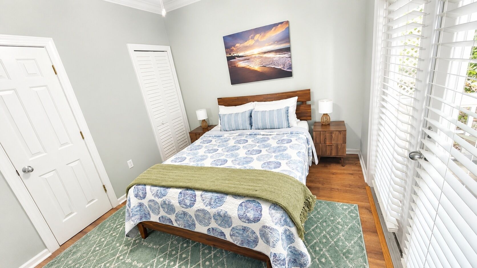 A bedroom with a bed, a nightstand, a lamp, a window with blinds, and a door.