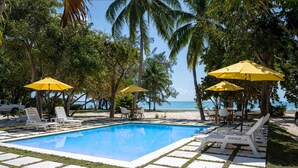 A pool area with lounge chairs and yellow umbrellas, surrounded by palm trees and a view of the beach.