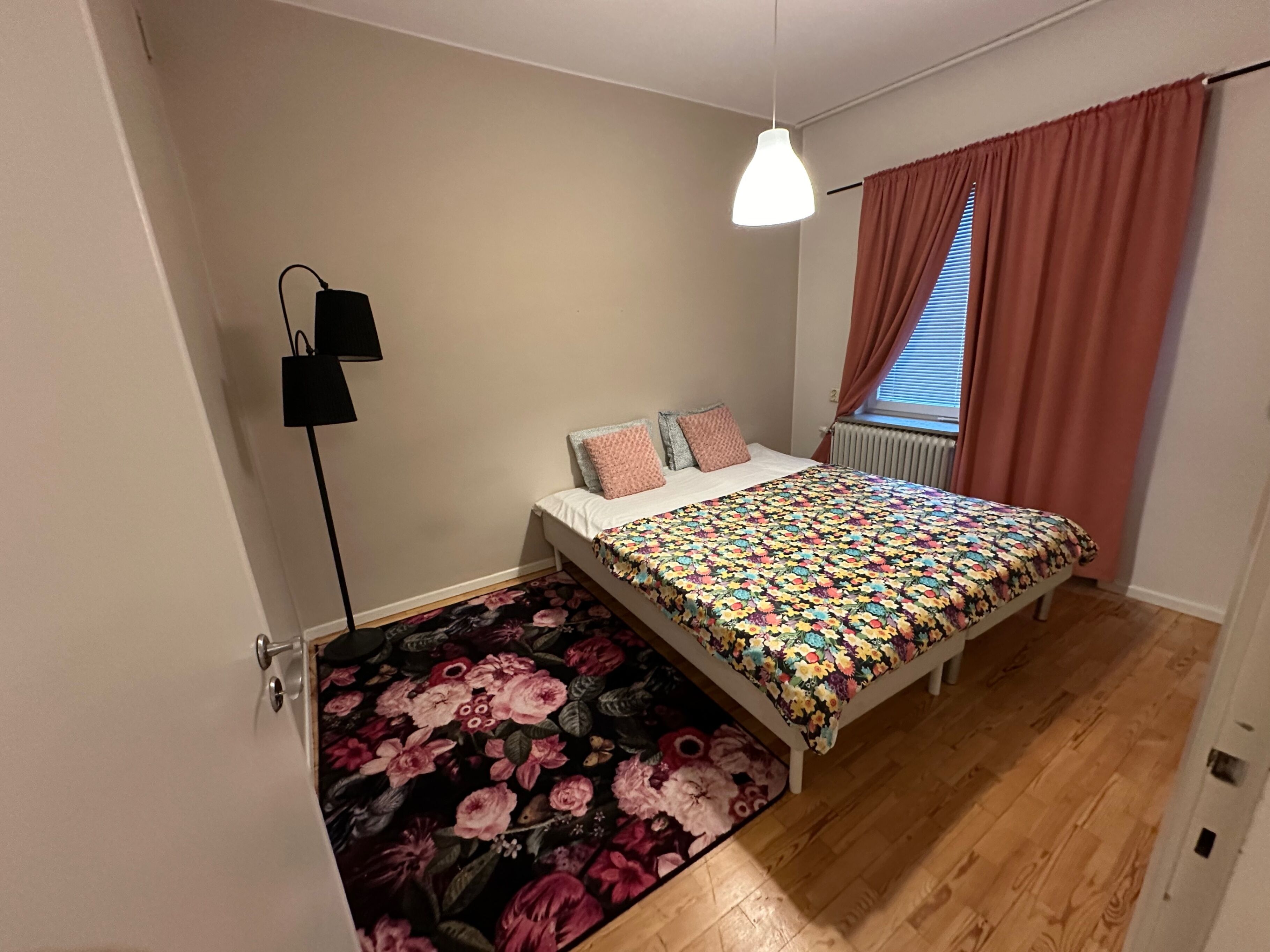 A bedroom with a bed, a floor lamp, a window with curtains, and a radiator.