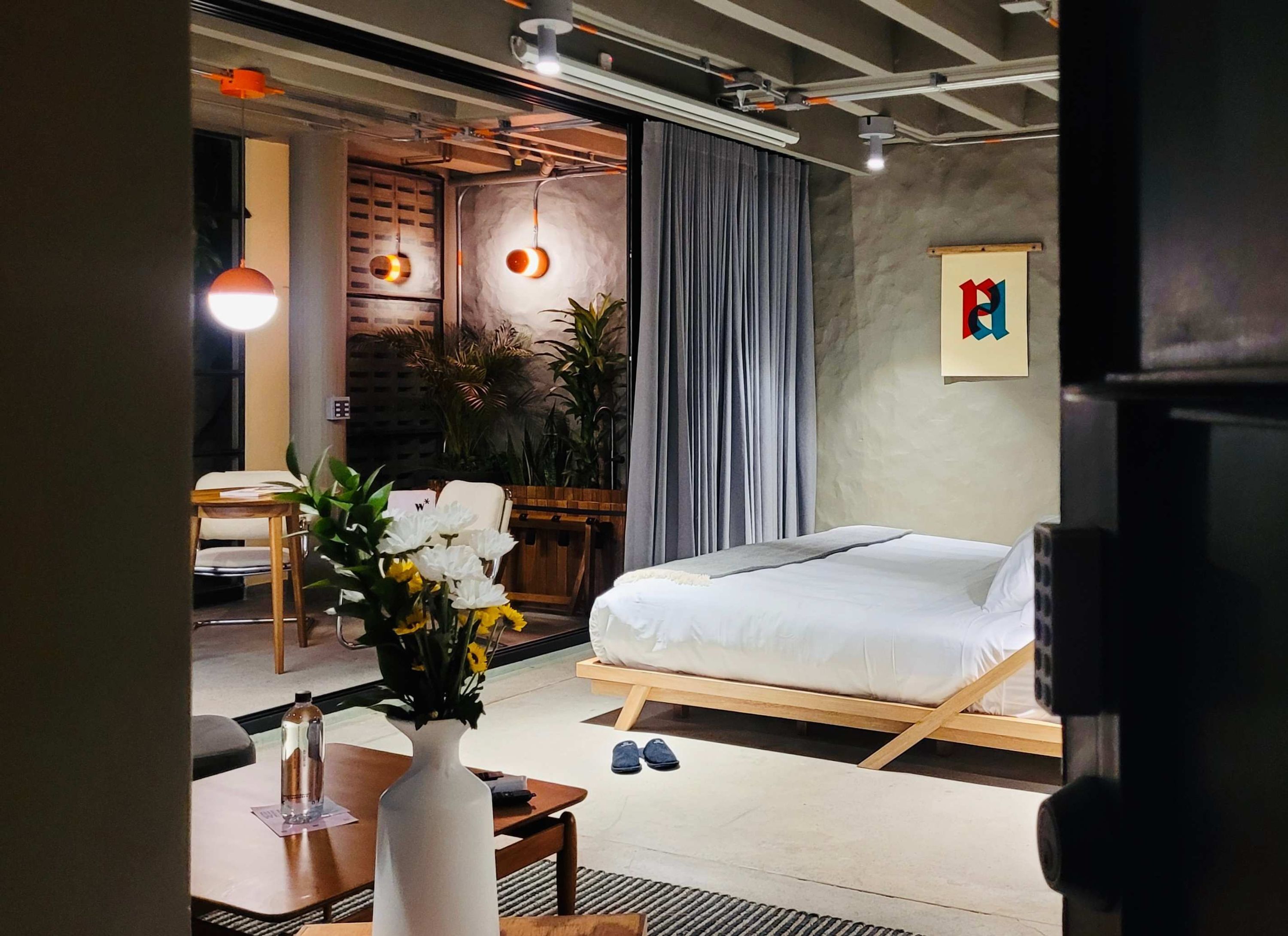 A modern bedroom with a bed, a desk, and a chair. There are plants and a painting on the wall.