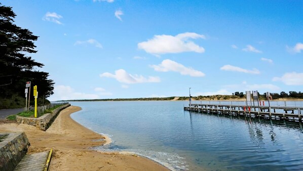 Une plage de sable avec une jetée en bois qui s’avance dans des eaux calmes, entourée de verdure et sous un ciel dégagé.