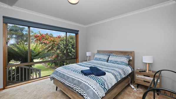 Ein Schlafzimmer mit großem Fenster, einem Bett mit gemusterten Betttextilien, einem Nachttisch mit Lampe und einem Sessel.