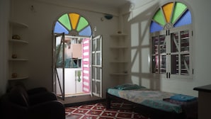 Une pièce comprenant un lit, une chaise, une étagère avec un bol, et une fenêtre avec des vitraux colorés.