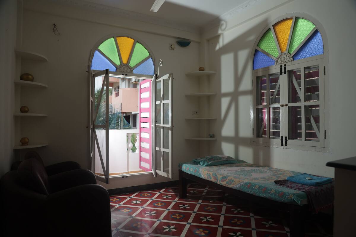 Een kamer met een bed, een stoel, een plankje met een schaal, en een raam met kleurrijke glaspanelen.