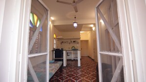 Un couloir étroit avec un ventilateur de plafond, un lit recouvert d’une courtepointe colorée et un coin cuisine équipé d’un réfrigérateur et d’étagères.